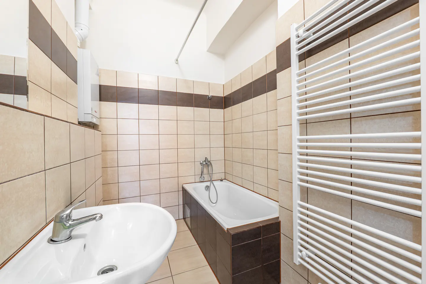 Bathroom with beige and brown tiled walls, a white sink, a bathtub with a handheld shower, and a white towel radiator.
