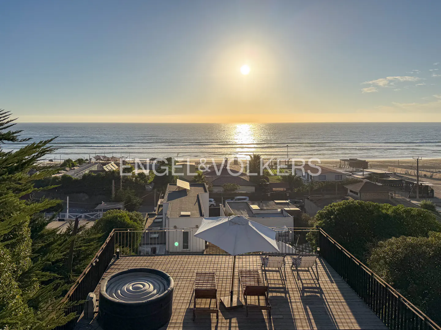 Rooftop view of ocean at sunset. Hot tub, chairs, and umbrella on wood deck. Houses and trees below.