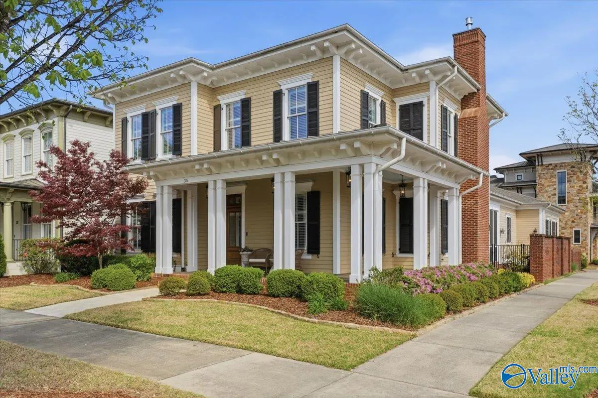 Two-story tan house with white trim, black shutters, and a brick chimney. A covered porch has white columns. Landscaped yard with a sidewalk.
