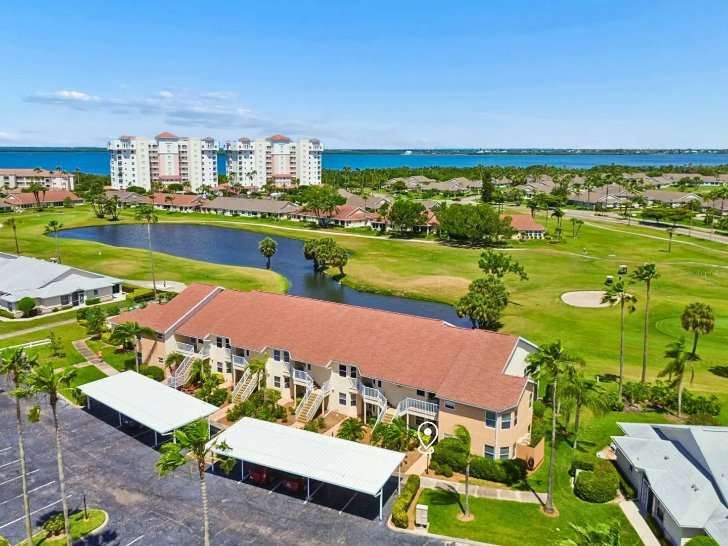 Aerial view of a tan condo building with a red tile roof, parking, and a golf course with a pond in the background.