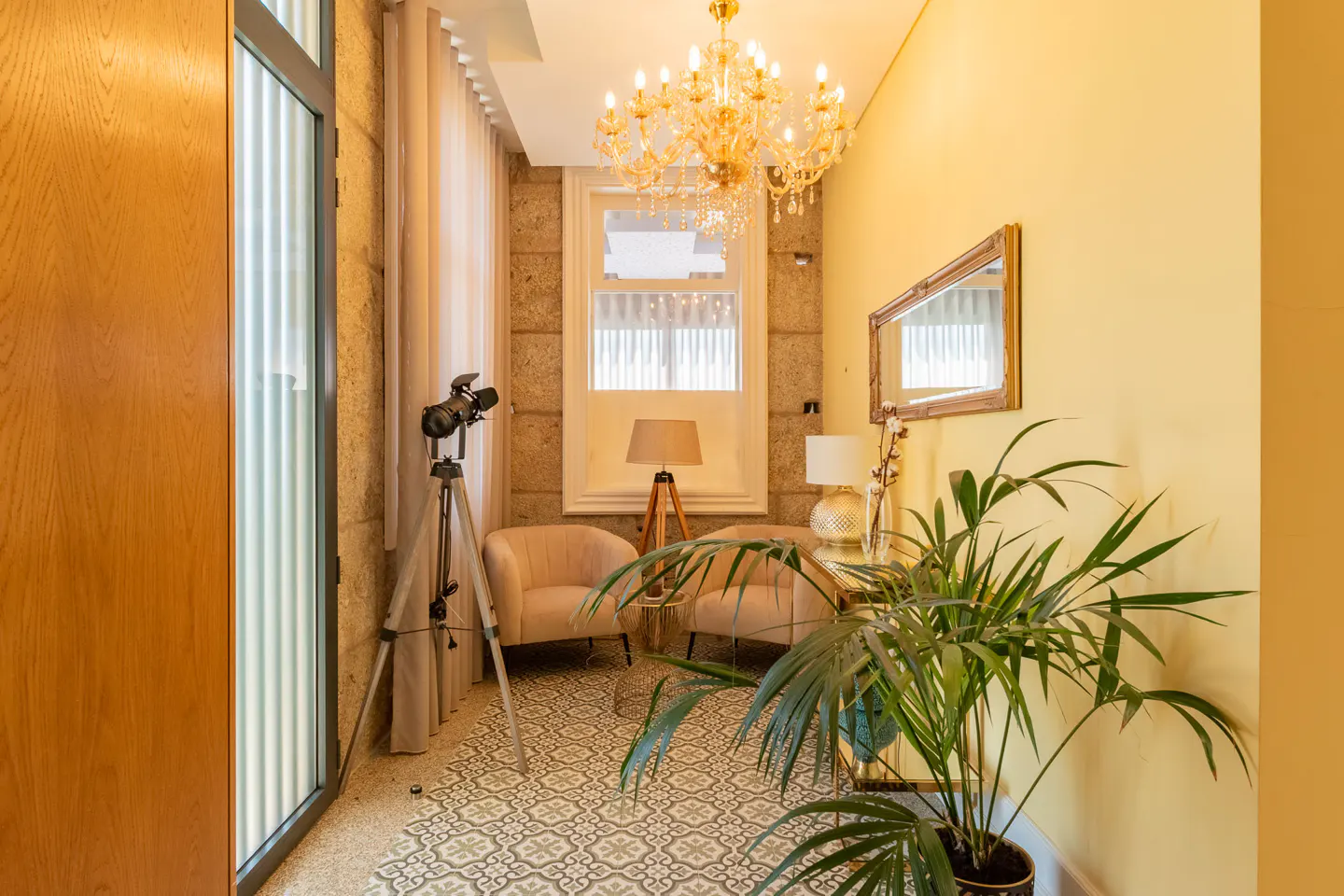 A bright room with yellow walls, a crystal chandelier, and two armchairs. A plant, mirror, and lamp decorate the space. A spotlight stands near a glass door.
