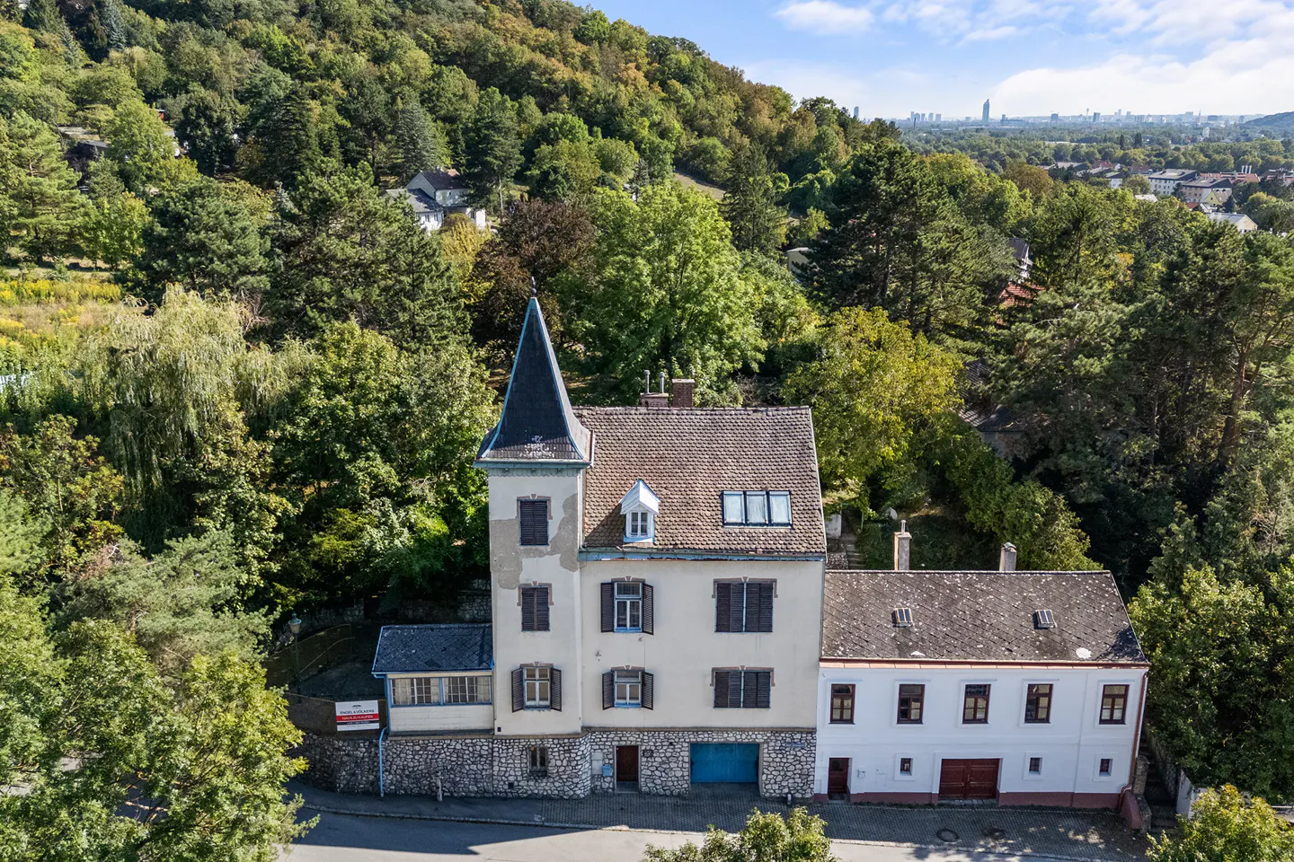 Aerial view of a beige house with a dark pointed roof, surrounded by green trees and a distant cityscape.