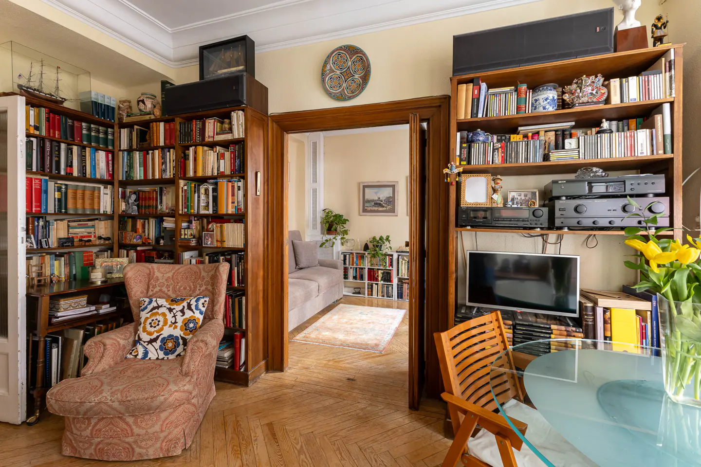 A cozy living room with floor-to-ceiling bookshelves, a patterned armchair, and a glimpse into another room with a sofa.