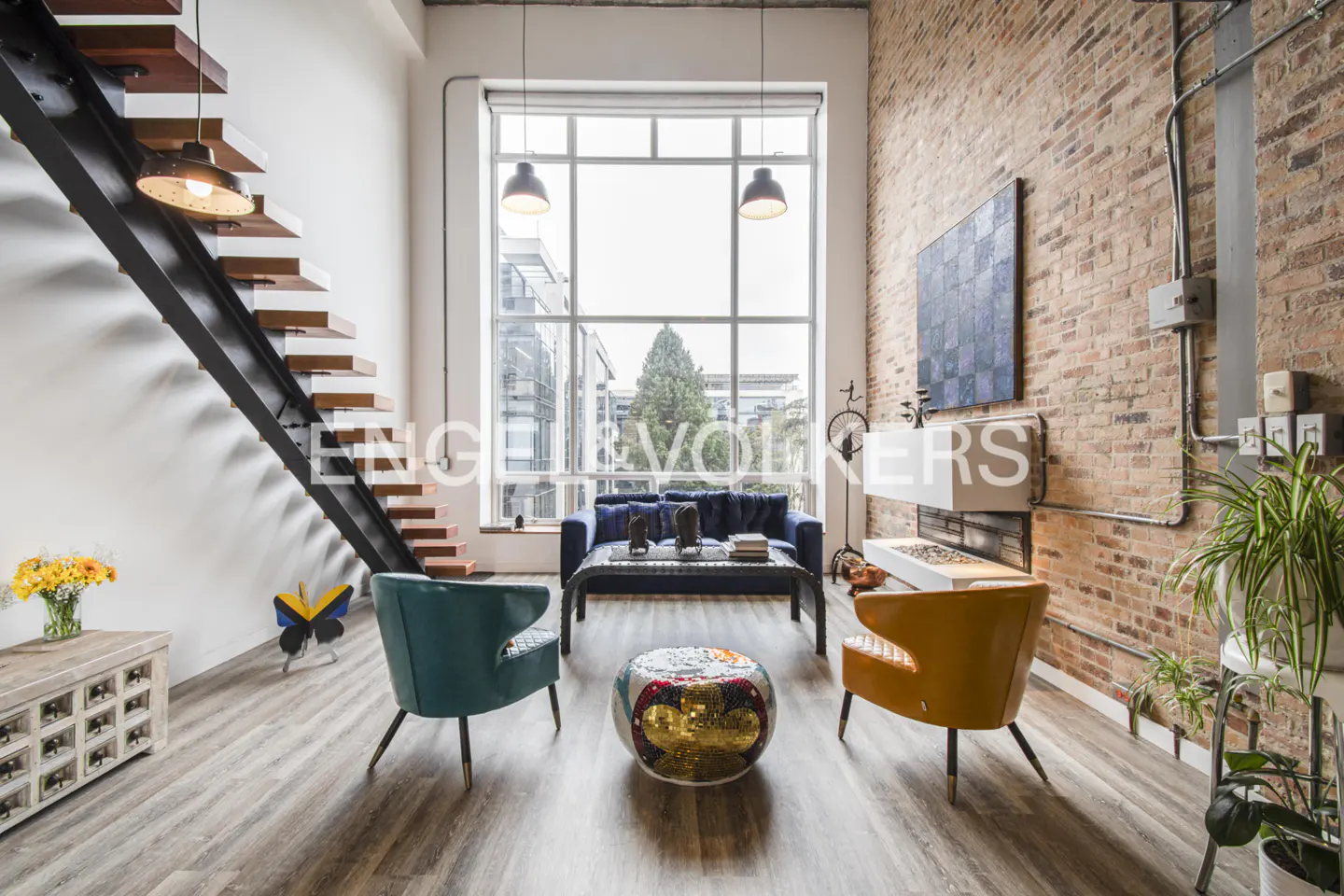 Loft apartment with exposed brick, large window, and modern furniture. A staircase is on the left, and a blue sofa is in the background.