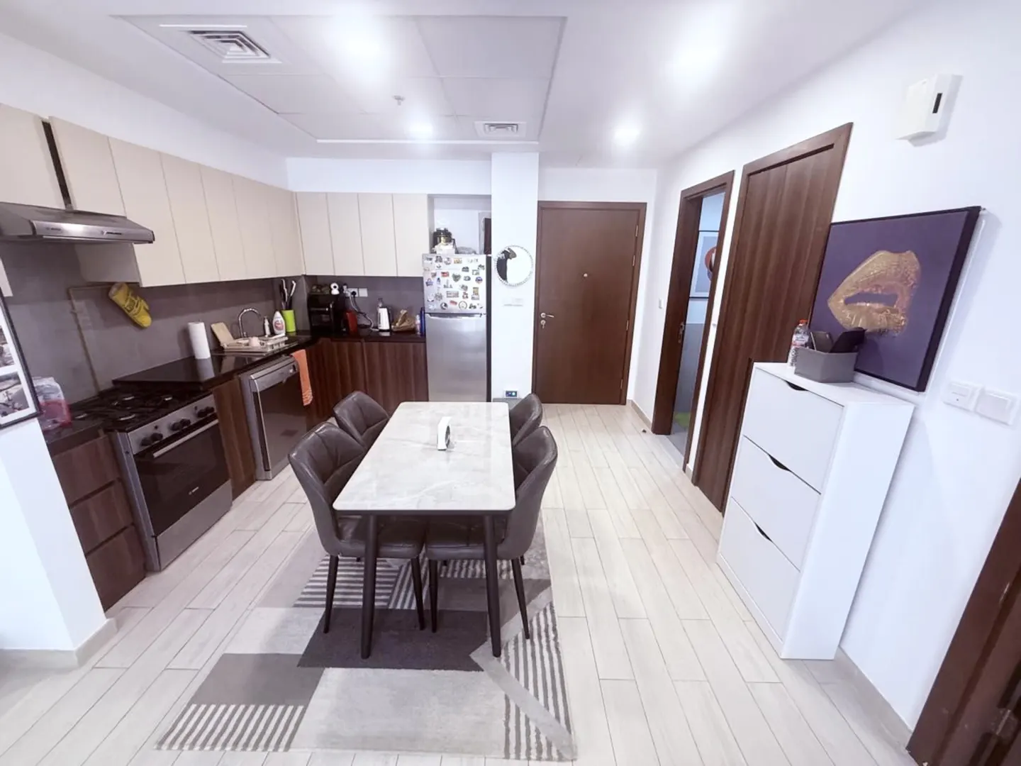 A modern apartment interior with a kitchen, dining area, and hallway. The kitchen features stainless steel appliances and dark wood cabinets. A marble table with six chairs sits on a striped rug.
