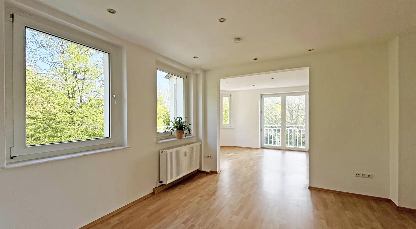 Bright, empty room with hardwood floors, white walls, and two windows overlooking green trees. A radiator sits beneath a window with a potted plant. A doorway leads to a balcony.