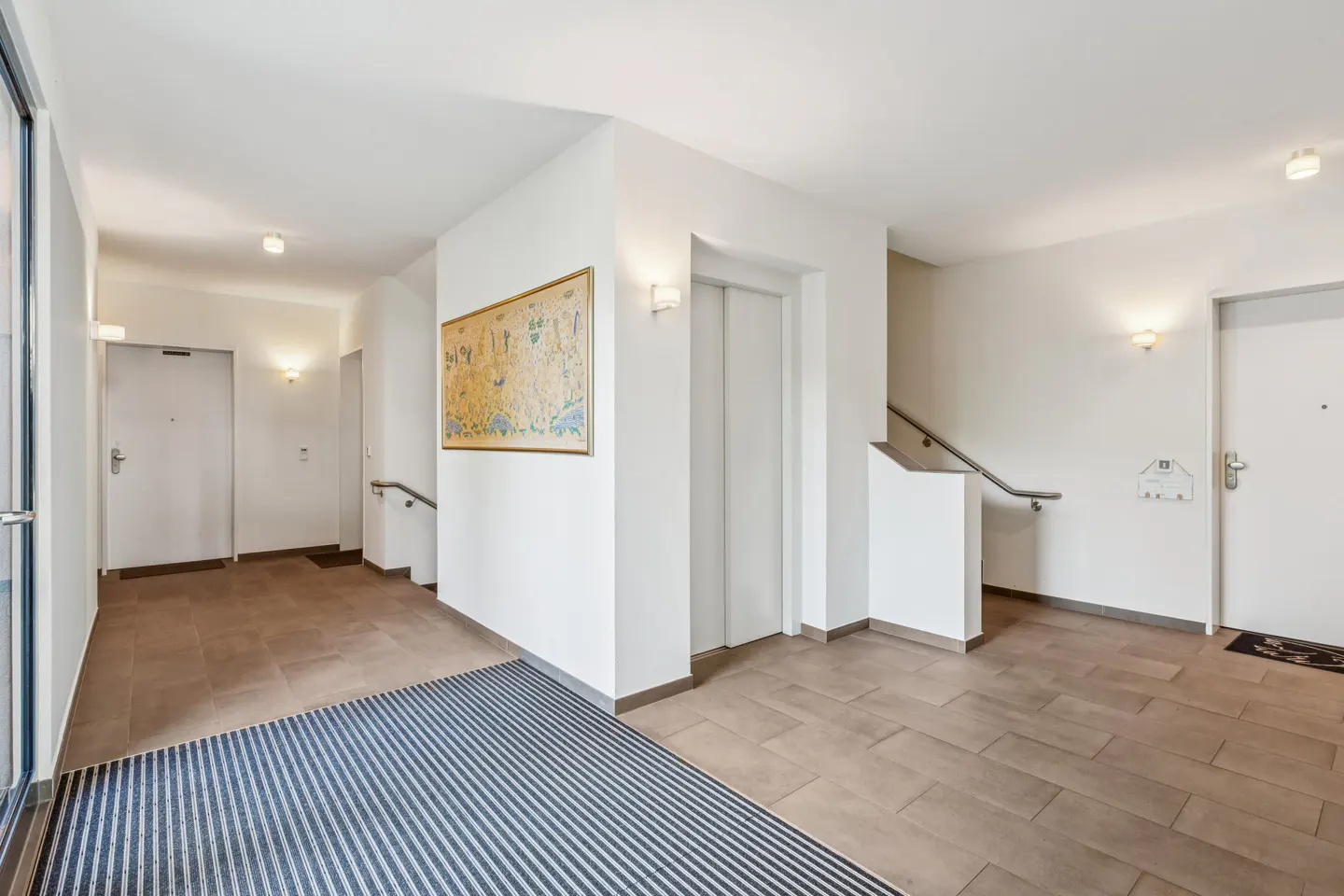 Apartment hallway with beige tile floor, white walls, elevator, stairs, and doors. A blue striped mat is in the foreground.