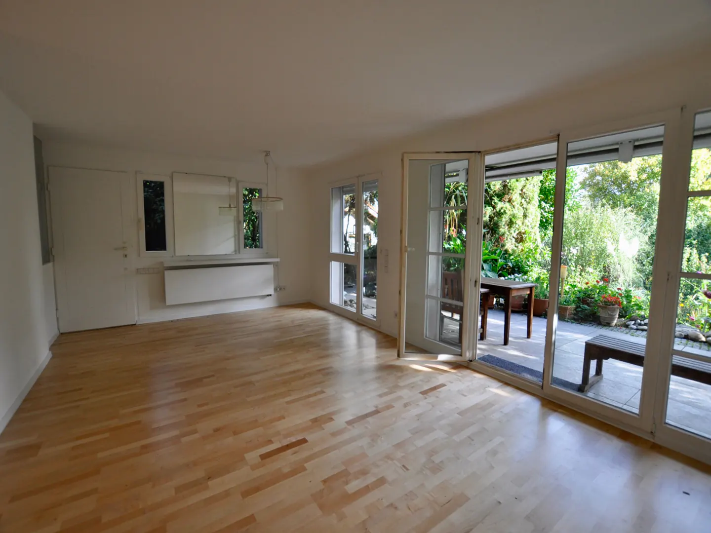 Bright, empty room with light wood floors and white walls. Glass doors open to a patio with greenery, table, and bench.