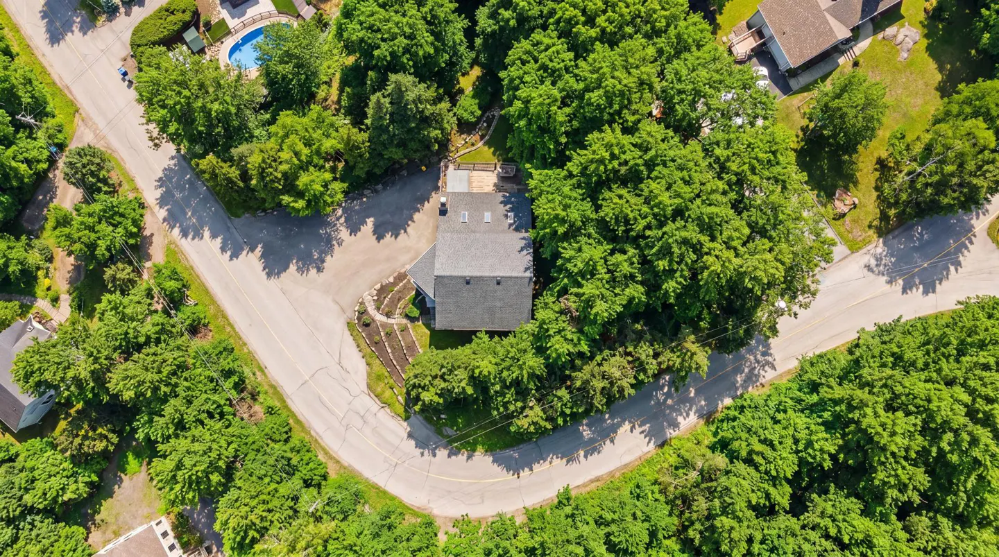 Aerial view of a gray roofed house surrounded by green trees and a winding road. A blue swimming pool is visible in the background.