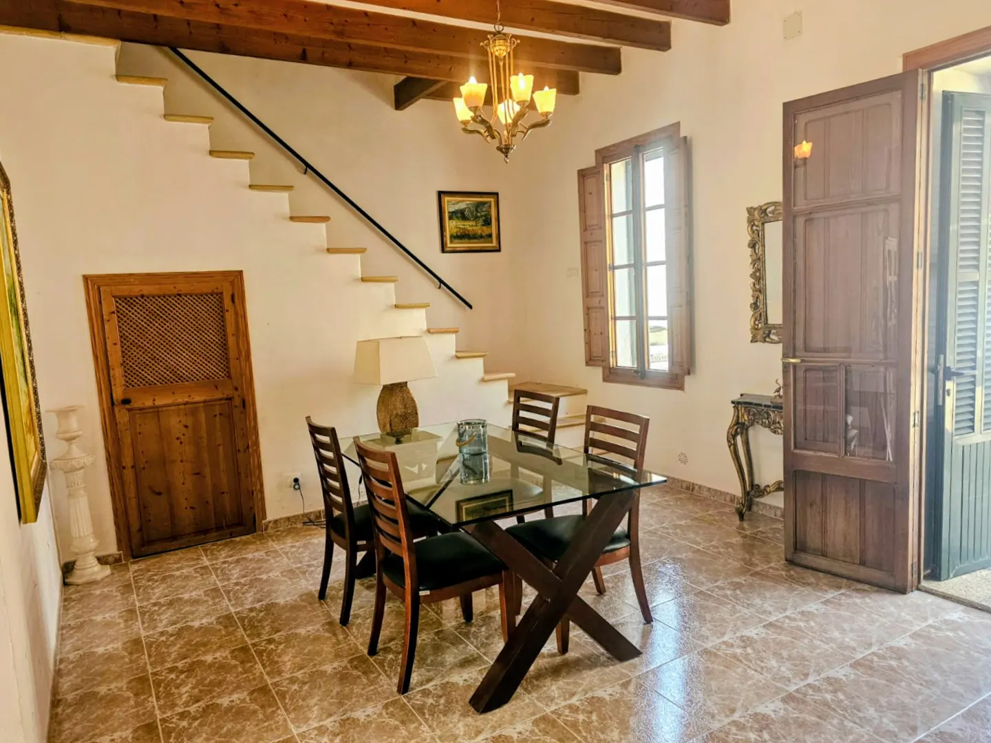 Bright dining room with glass table, wooden chairs, and open door. Stairs lead up to the second floor. Chandelier hangs from the wooden beamed ceiling.