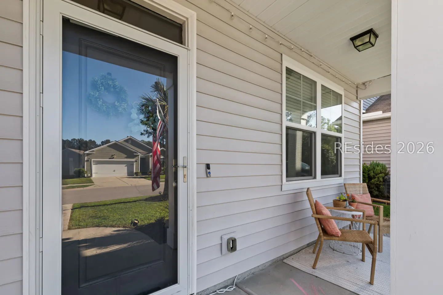 Exterior view of a house with a white door reflecting a house across the street and an American flag. Two chairs sit on the porch.