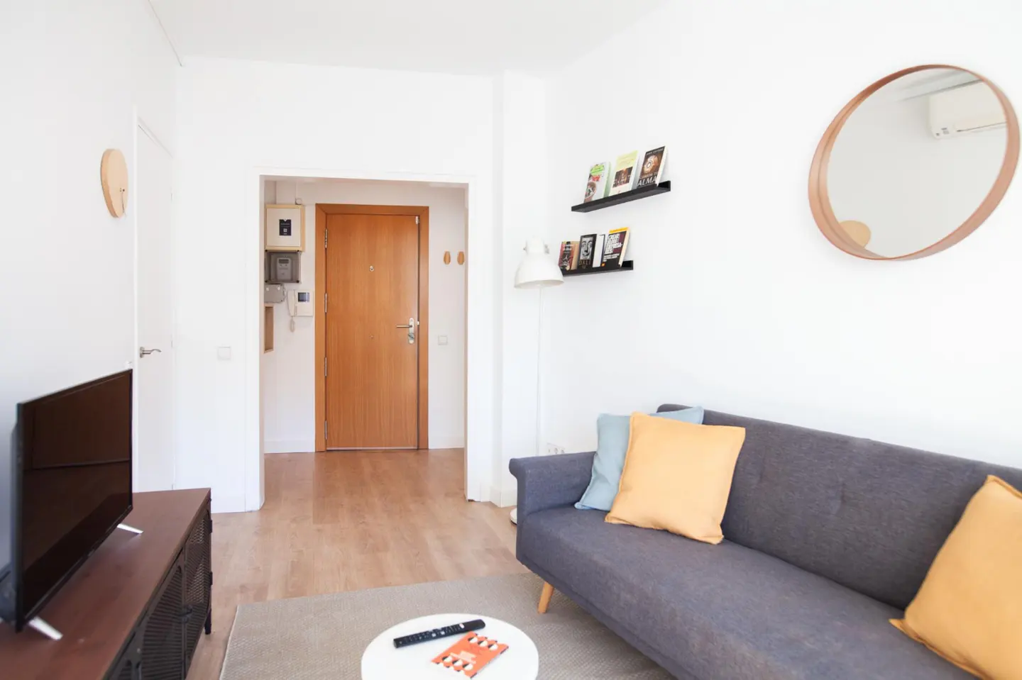 Living room with gray sofa, yellow and blue pillows, round mirror, and wooden door. Bookshelves on the white wall.