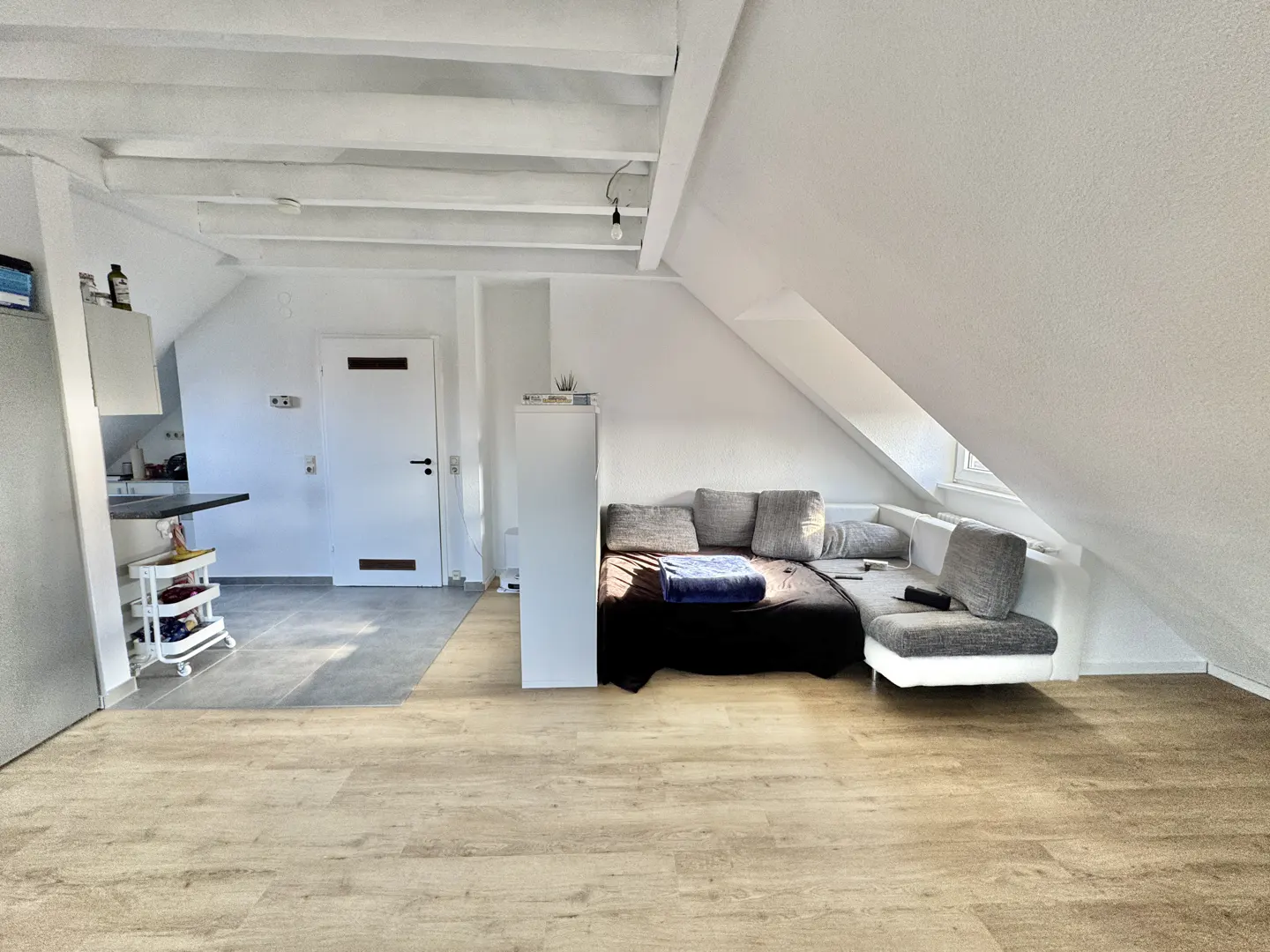 Attic apartment with white walls, exposed beams, and light wood floors. A gray sectional sofa sits under a window. A small kitchen area is visible.