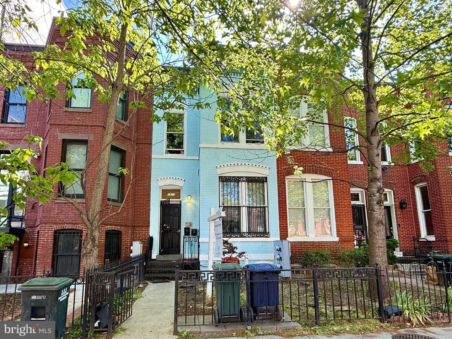 Row houses in a city neighborhood. The center house is light blue, flanked by red brick houses. A black iron fence lines the front.