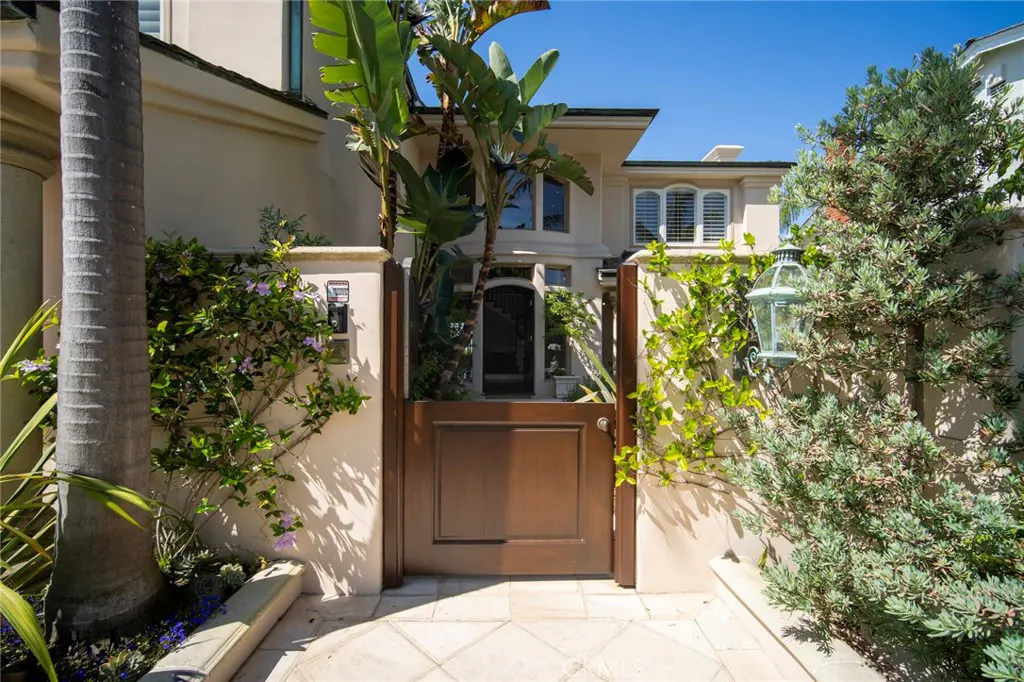 Exterior view of a tan house with a brown gate, lush greenery, and a stone walkway.