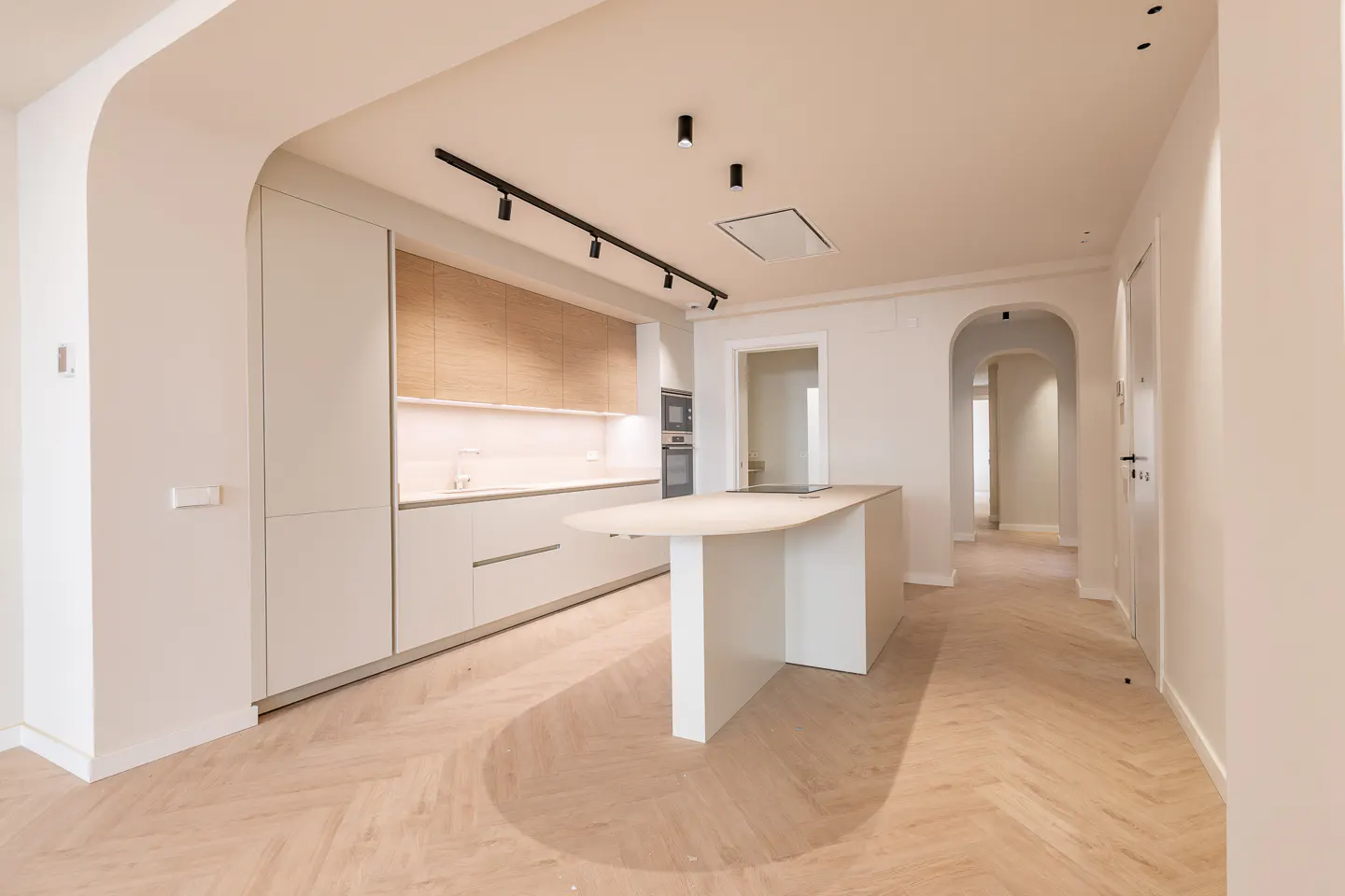 Bright, modern kitchen with white cabinets, wood accents, and a large island. Herringbone wood floors and arched doorways add architectural detail.