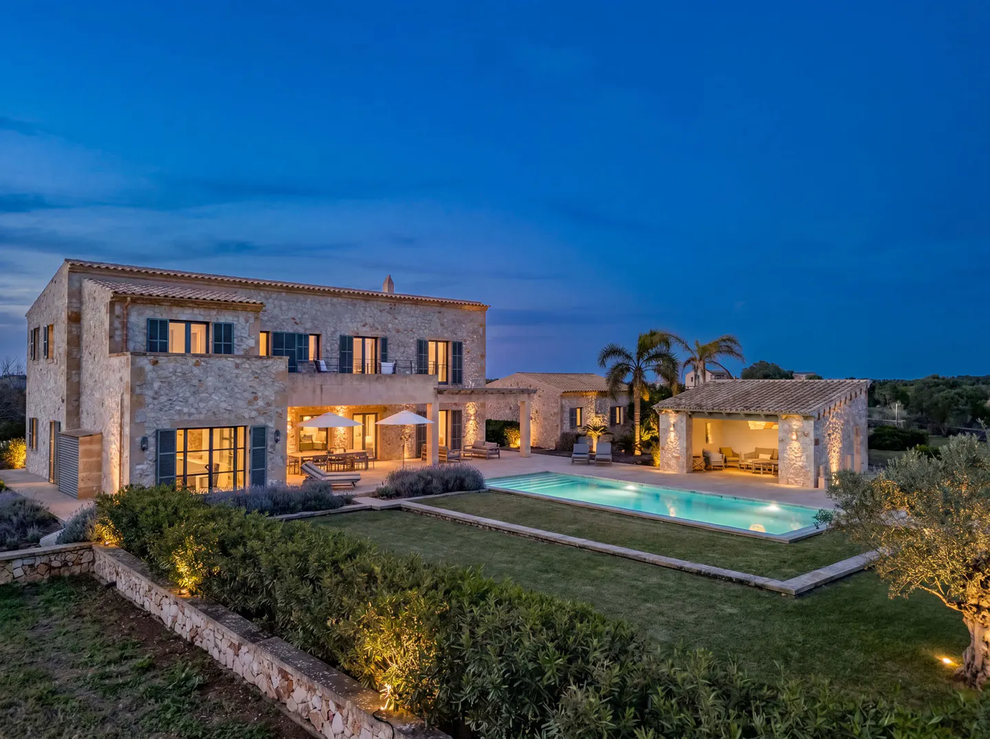 A stone house with a pool at dusk. The house has dark shutters and lights on inside. The pool is lit and surrounded by grass.