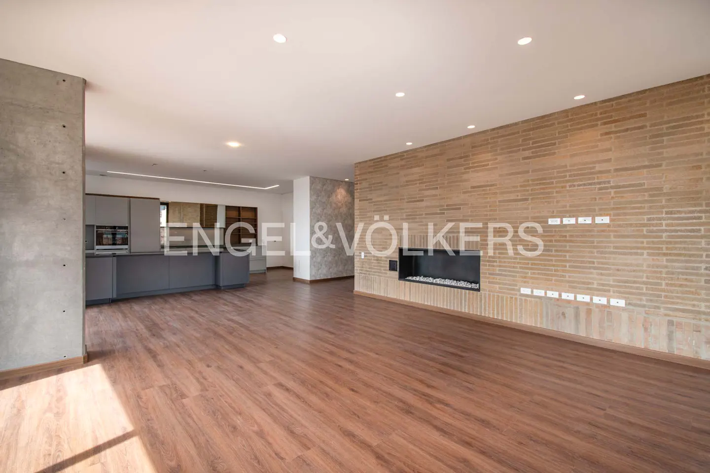 A spacious living room with wood floors, a brick wall with a fireplace, and a gray kitchen in the background.