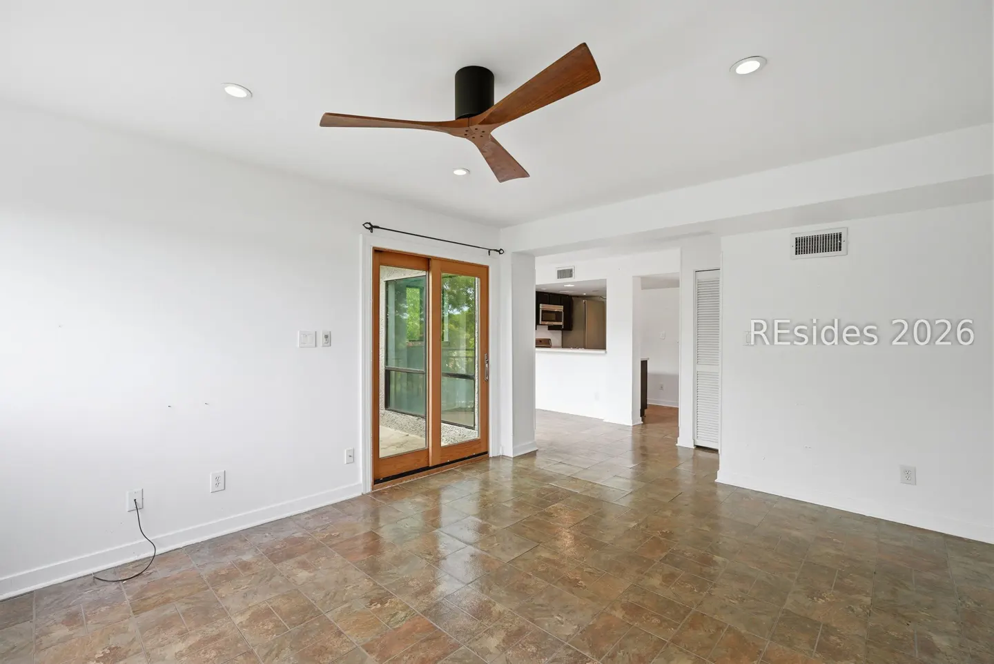 Bright, empty room with tile floor, white walls, and a modern ceiling fan. Sliding glass doors lead to the outside. Kitchen visible in the background.
