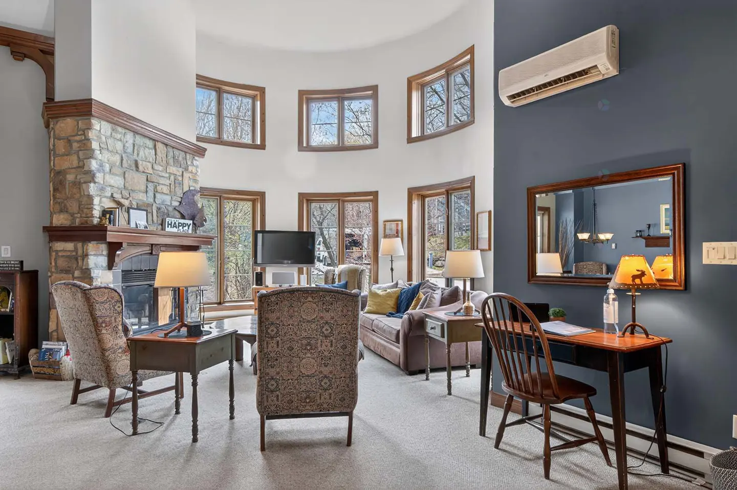 Living room with stone fireplace, curved windows, brown sofa, patterned chairs, and a desk against a blue wall.