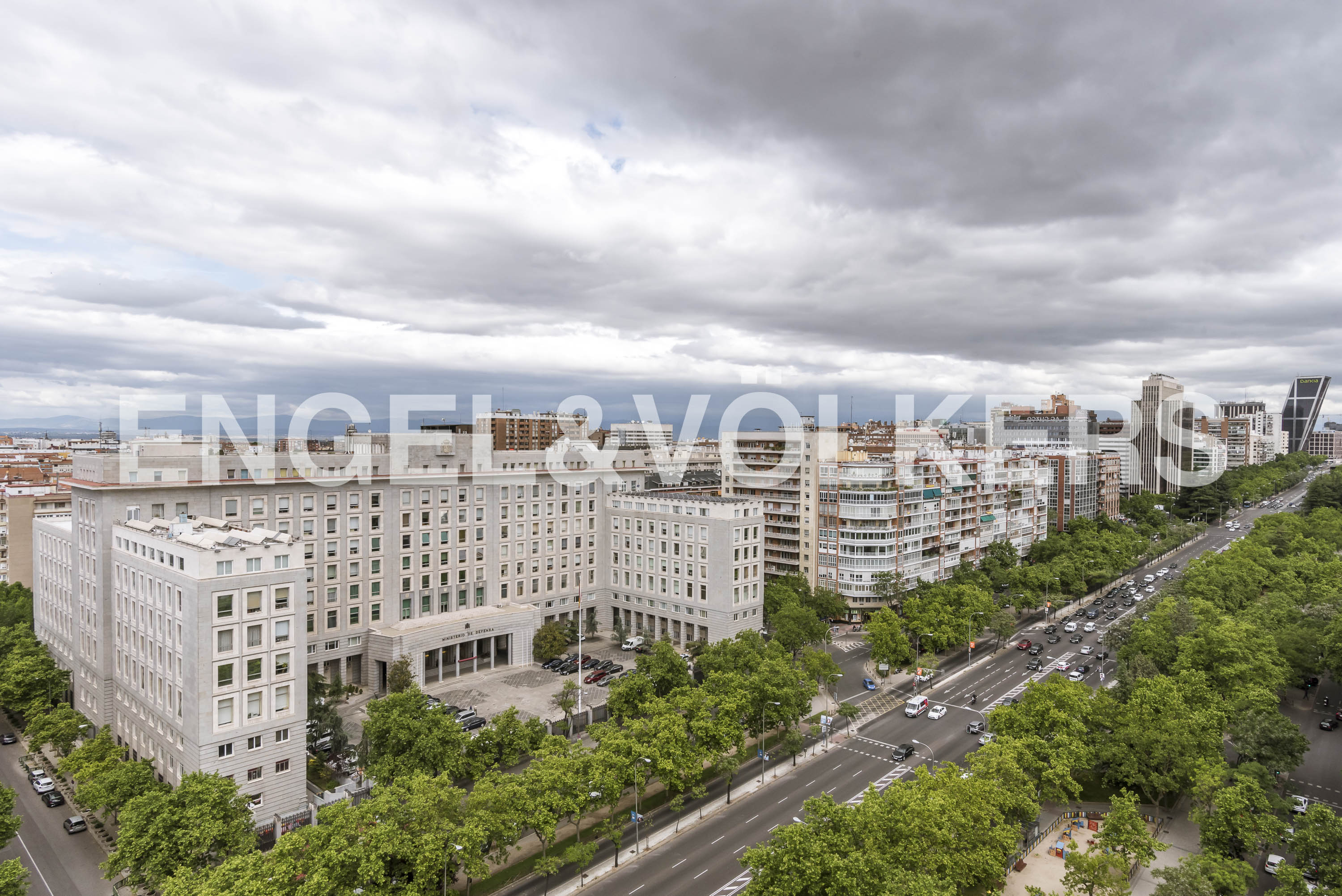High-angle view of a large grey office building in a city, next to a tree-lined street with traffic under a cloudy sky.
