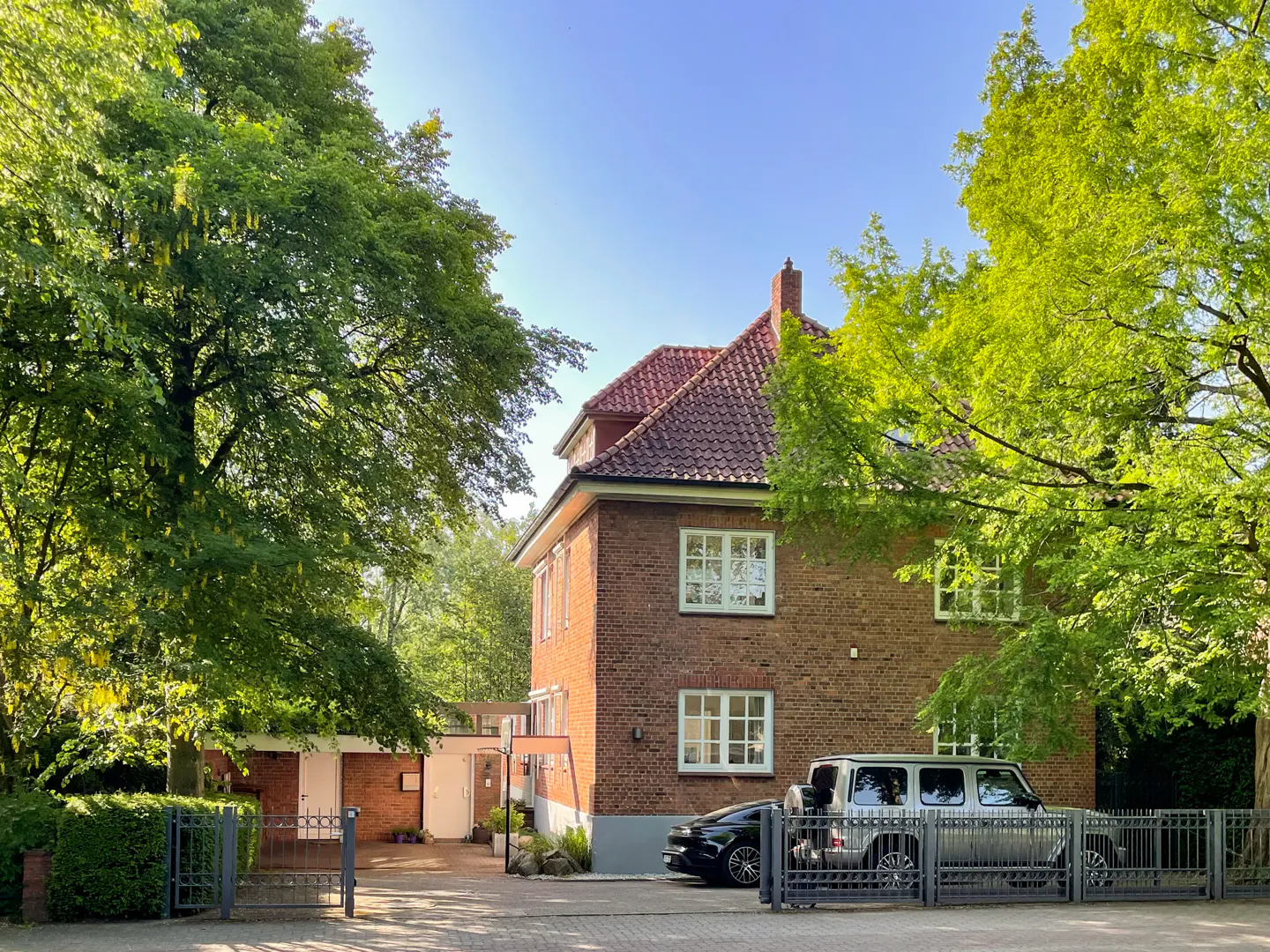 A two-story brick house with a red tile roof, surrounded by green trees, and two cars parked behind a gray metal fence.