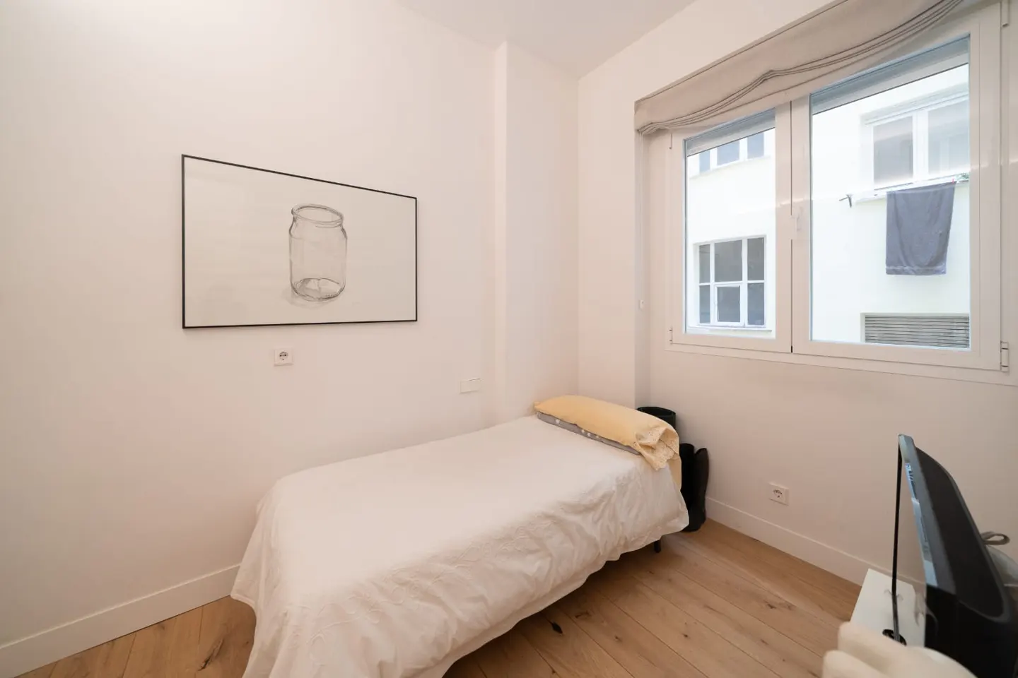A small bedroom with a single bed, white walls, and a window with a gray shade. A drawing of a jar hangs on the wall.
