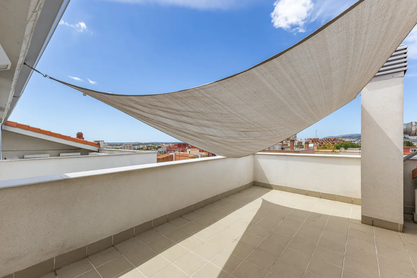 A sun-drenched balcony with beige tiles and a white canvas shade. City buildings are visible in the background under a blue sky.