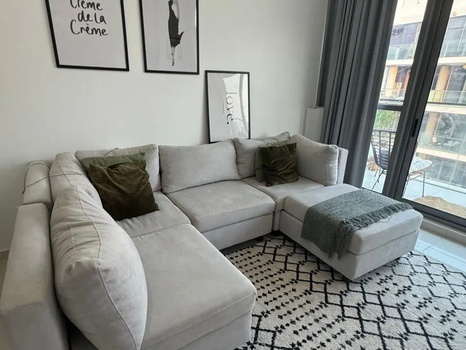 Living room with a large, light gray sectional sofa, olive green pillows, and a black and white patterned rug. Art on the wall and a balcony view.