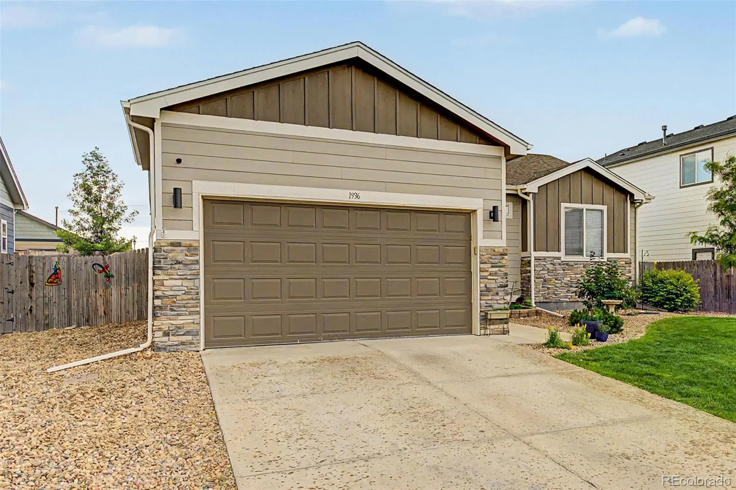 Exterior view of a tan and brown home with a two-car garage, stone accents, a concrete driveway, and a fenced yard.