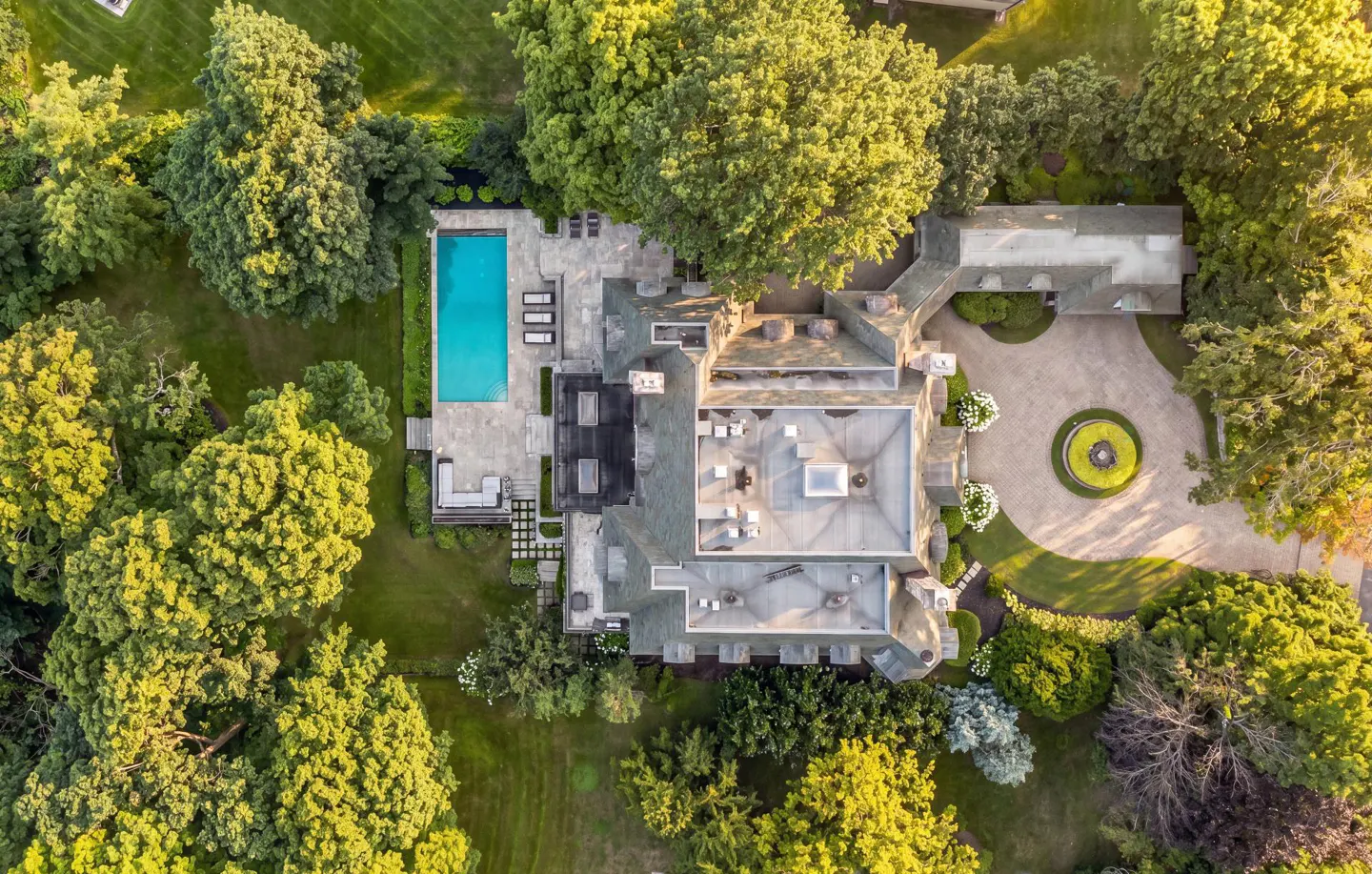 Aerial view of a large, gray stone house with a pool, circular driveway, and lush green trees surrounding the property.