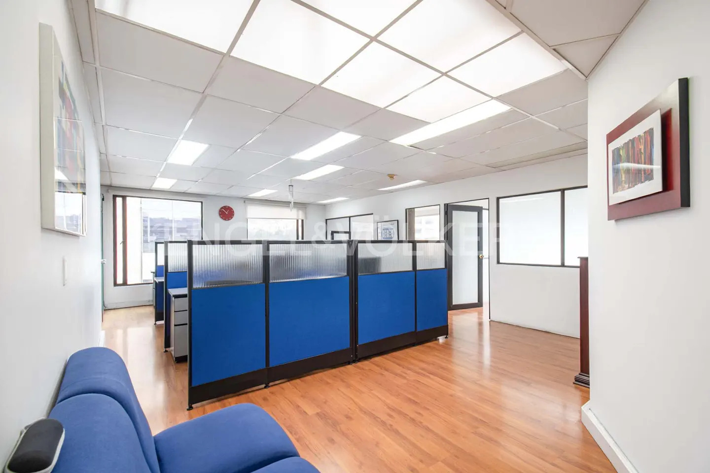 Office space with blue cubicles, wood floors, and white walls. Windows provide natural light. Artwork hangs on the walls.
