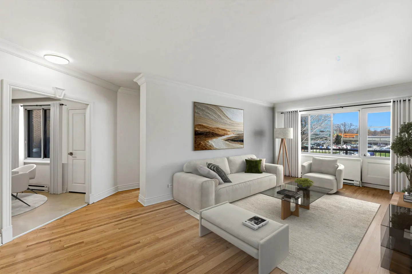 Bright living room with hardwood floors, white sofa, chair, and bench. Large windows offer a view of the outdoors.