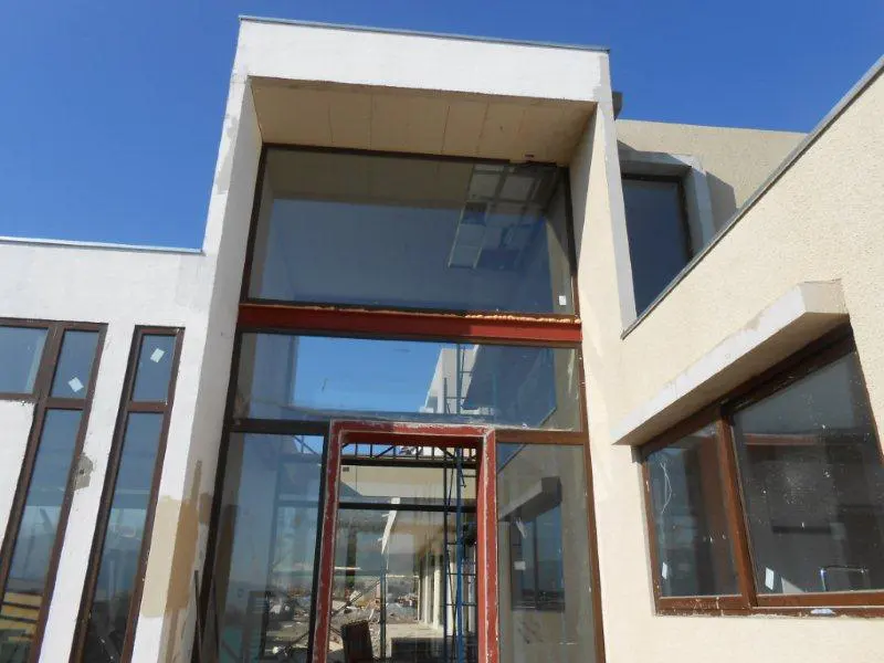 Exterior view of a modern building under construction with large windows and a red door frame.