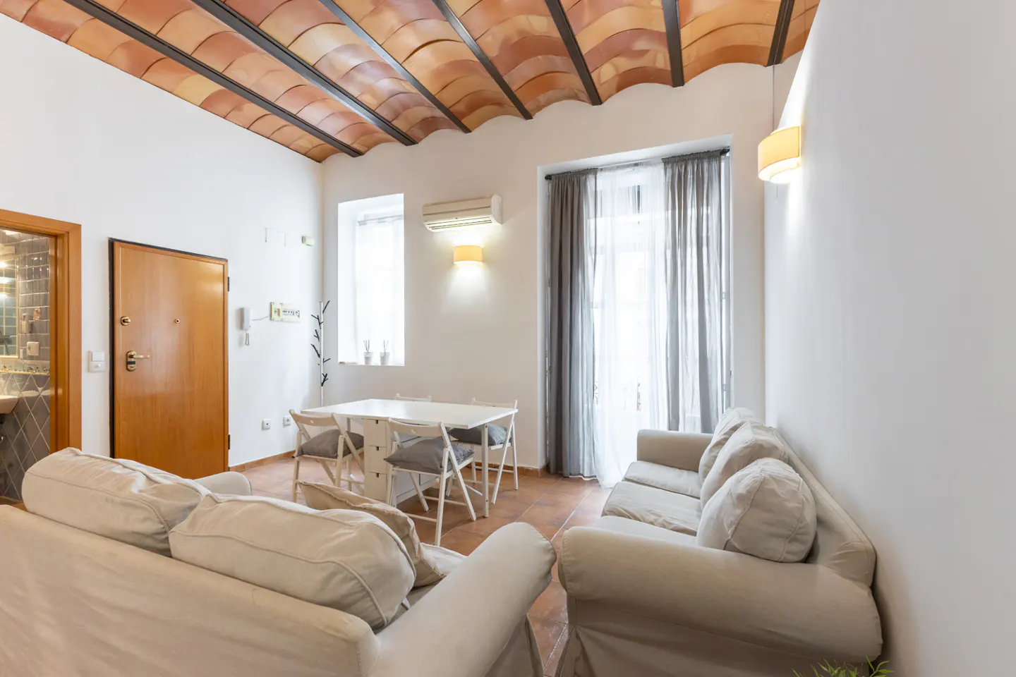 Bright living room with white walls, terracotta tile floor, and a vaulted tile ceiling. A white sofa and dining set furnish the space.