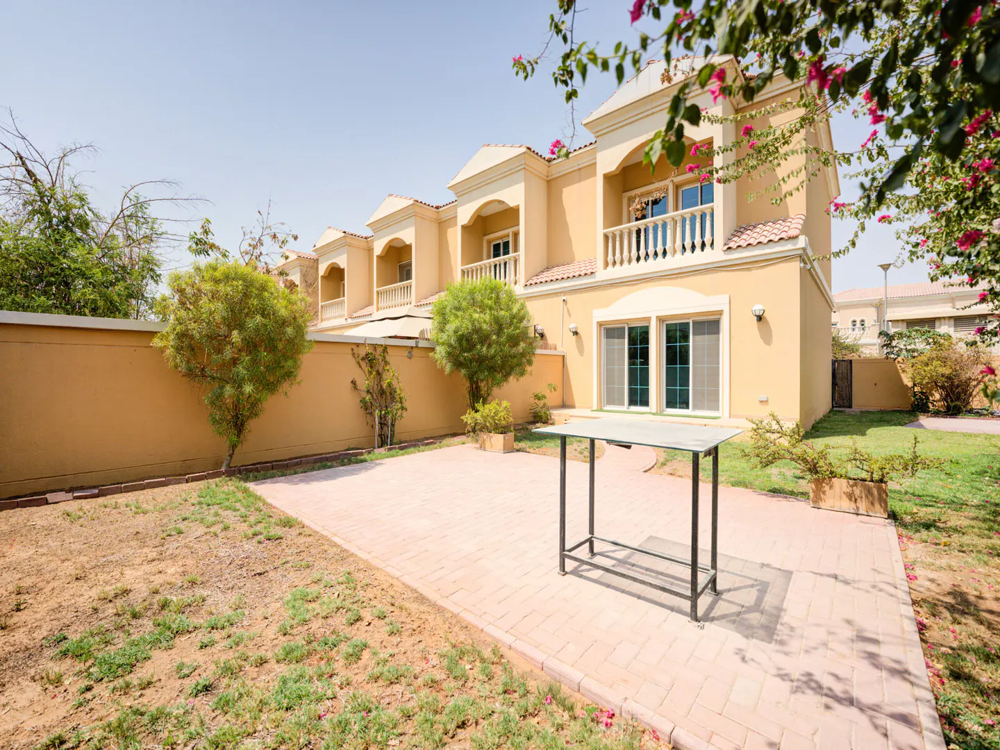 Backyard view of a two-story, tan townhouse with a patio, table, and green trees.