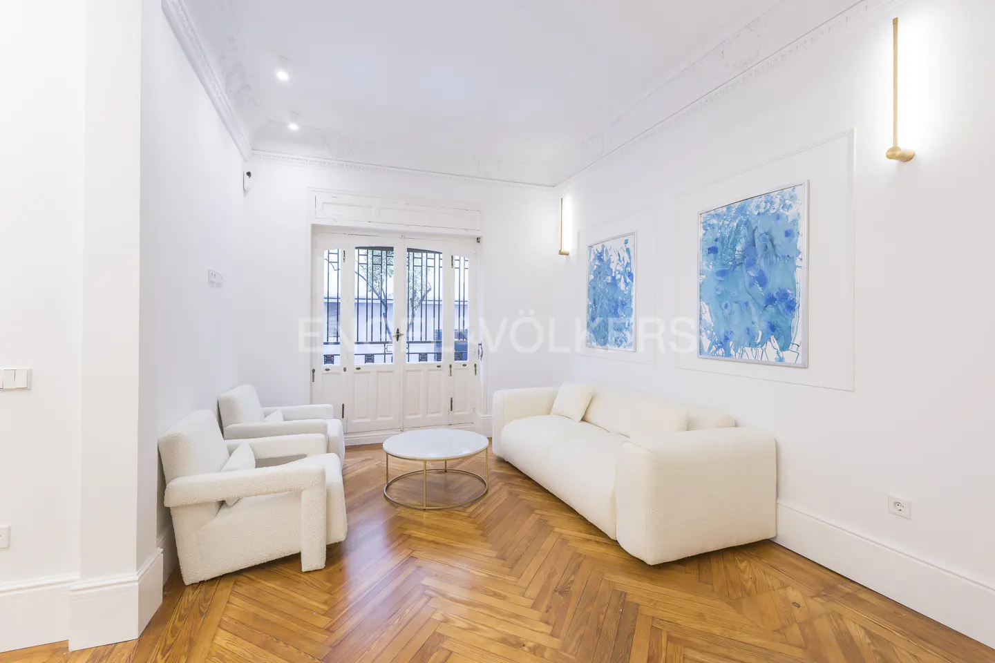 Bright living room with white walls, herringbone wood floor, white sofa and chairs, blue art, and white doors to a balcony.