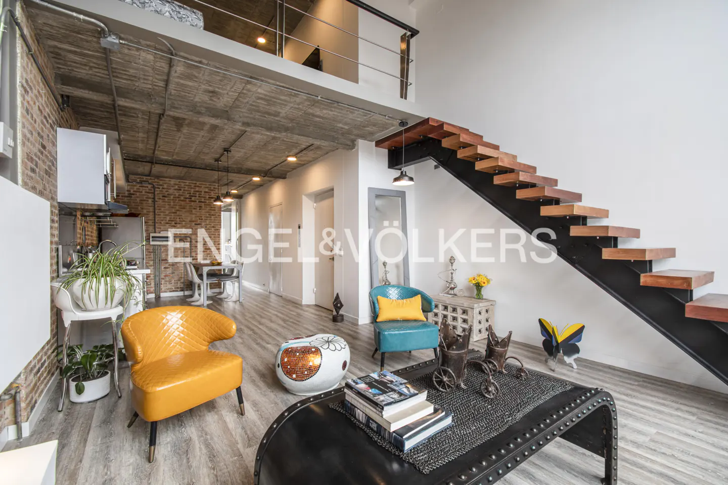 A modern loft interior with exposed brick, concrete ceiling, and floating wood stairs. Furnished with colorful chairs and a black coffee table.