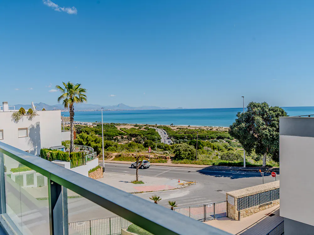 View from a balcony overlooking a road, beach, and ocean under a clear blue sky.