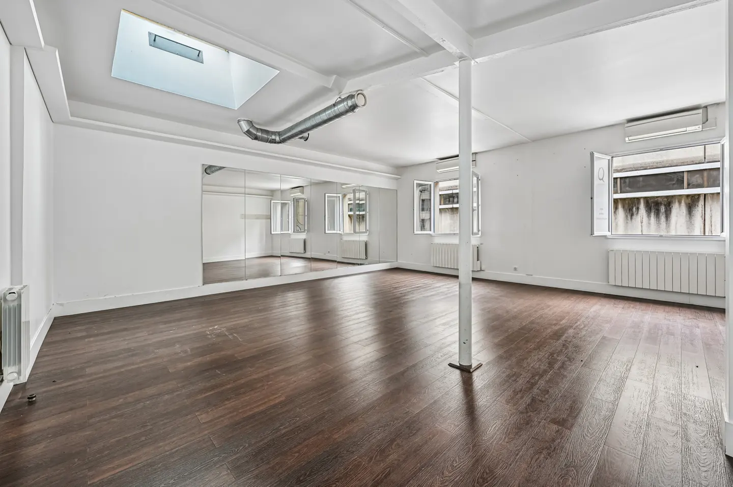 Bright, empty studio with dark wood floors, white walls, a wall mirror, and two windows. A skylight and metal duct are on the ceiling.