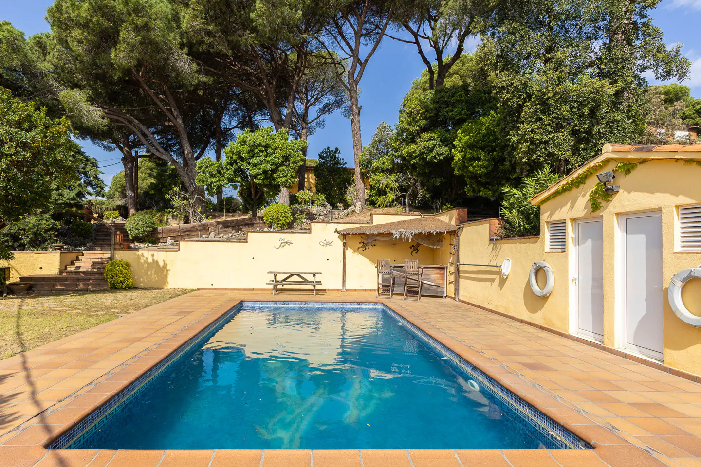 Rectangular blue pool with terracotta tile surround. Yellow building with white doors and life preservers. Trees and blue sky in background.