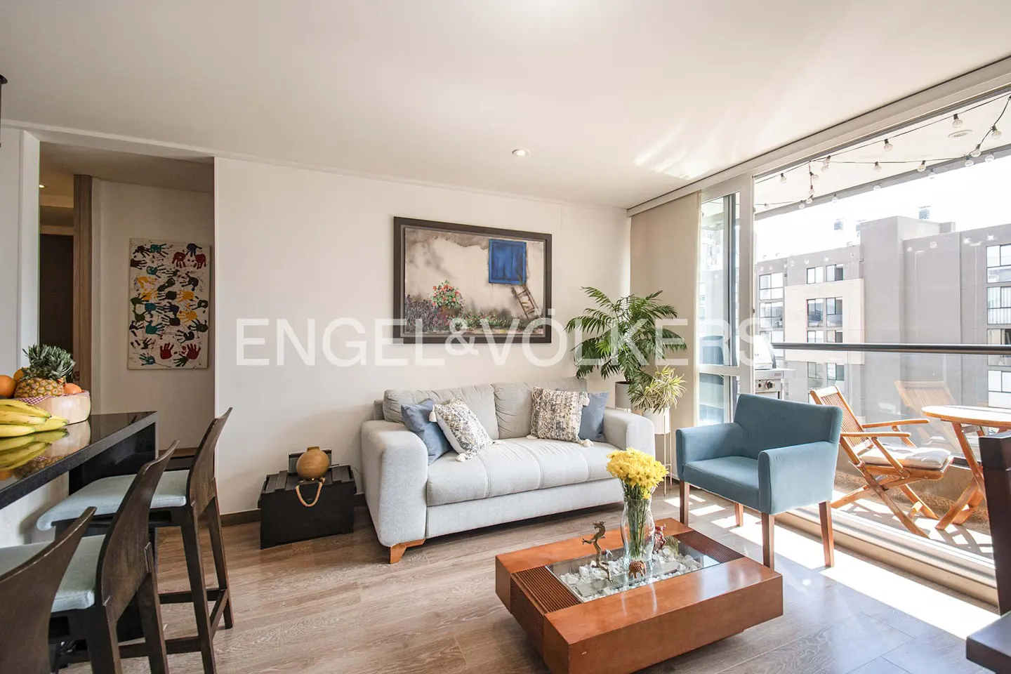 Bright living room with a gray sofa, blue chair, wood coffee table, and balcony with outdoor seating. Artwork adorns the walls.