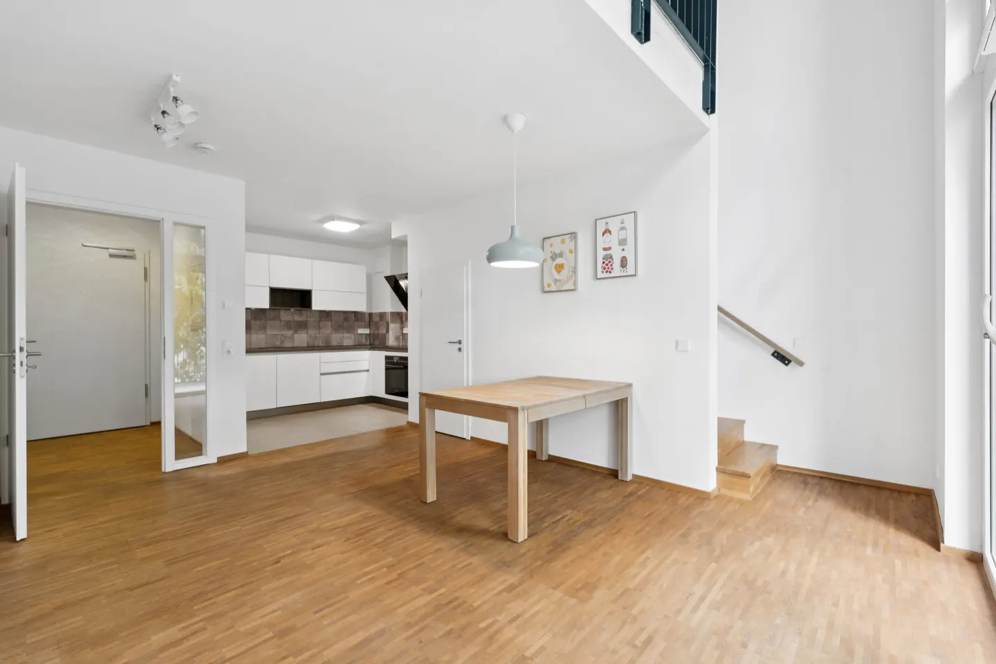 Bright, modern apartment interior with wood floors, white walls, and a wooden table. A kitchen is visible in the background.