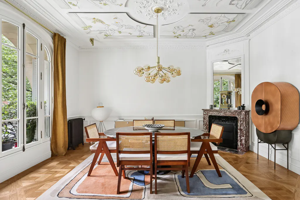 Elegant dining room with a round table, cane chairs, marble fireplace, and a modern gold chandelier under a painted ceiling.