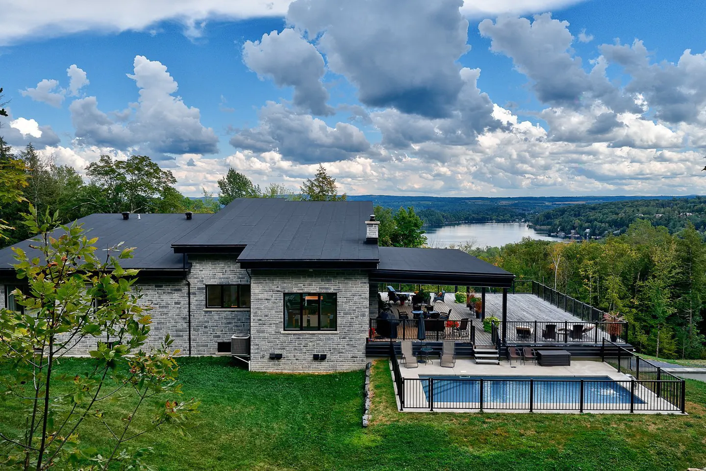 A modern stone house with a black roof, deck, and pool overlooks a lake surrounded by trees under a cloudy sky.