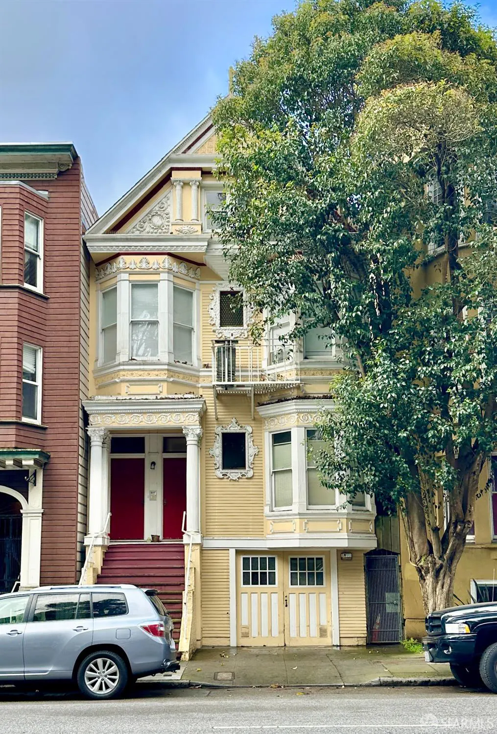 Street view of a yellow, multi-story Victorian house with red doors and a large tree in front. Cars are parked on the street.