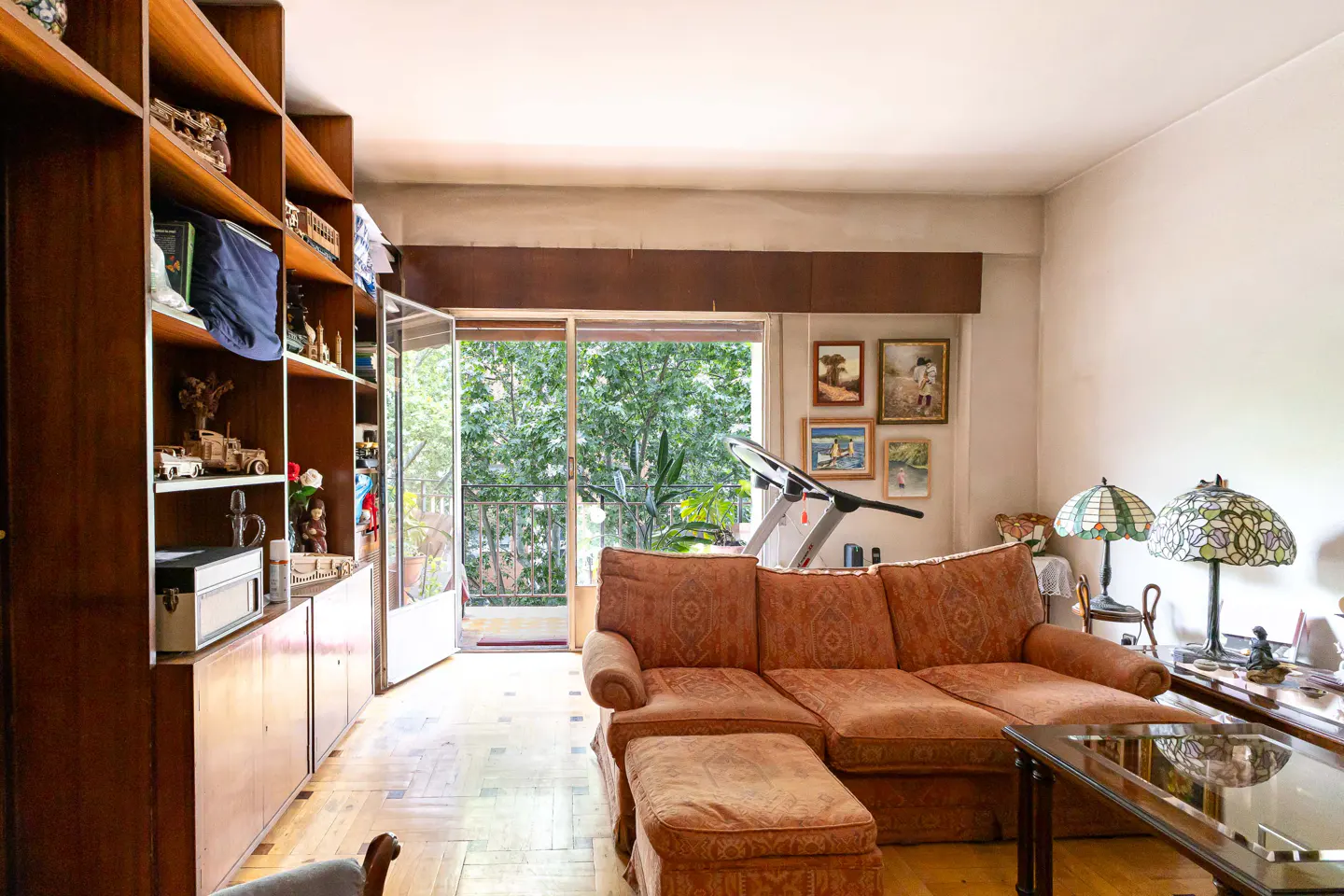 Living room with a brown patterned sofa, glass coffee table, and a bookshelf filled with items. Balcony doors open to a view of green trees.
