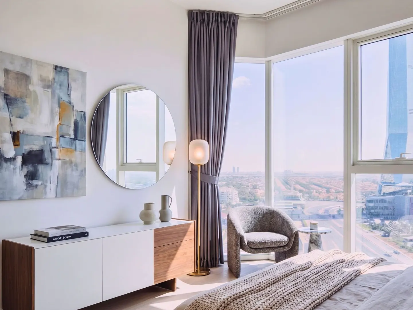 Bright bedroom with a large window, grey curtains, a round mirror, and a modern white and wood dresser. A grey chair sits near the window.