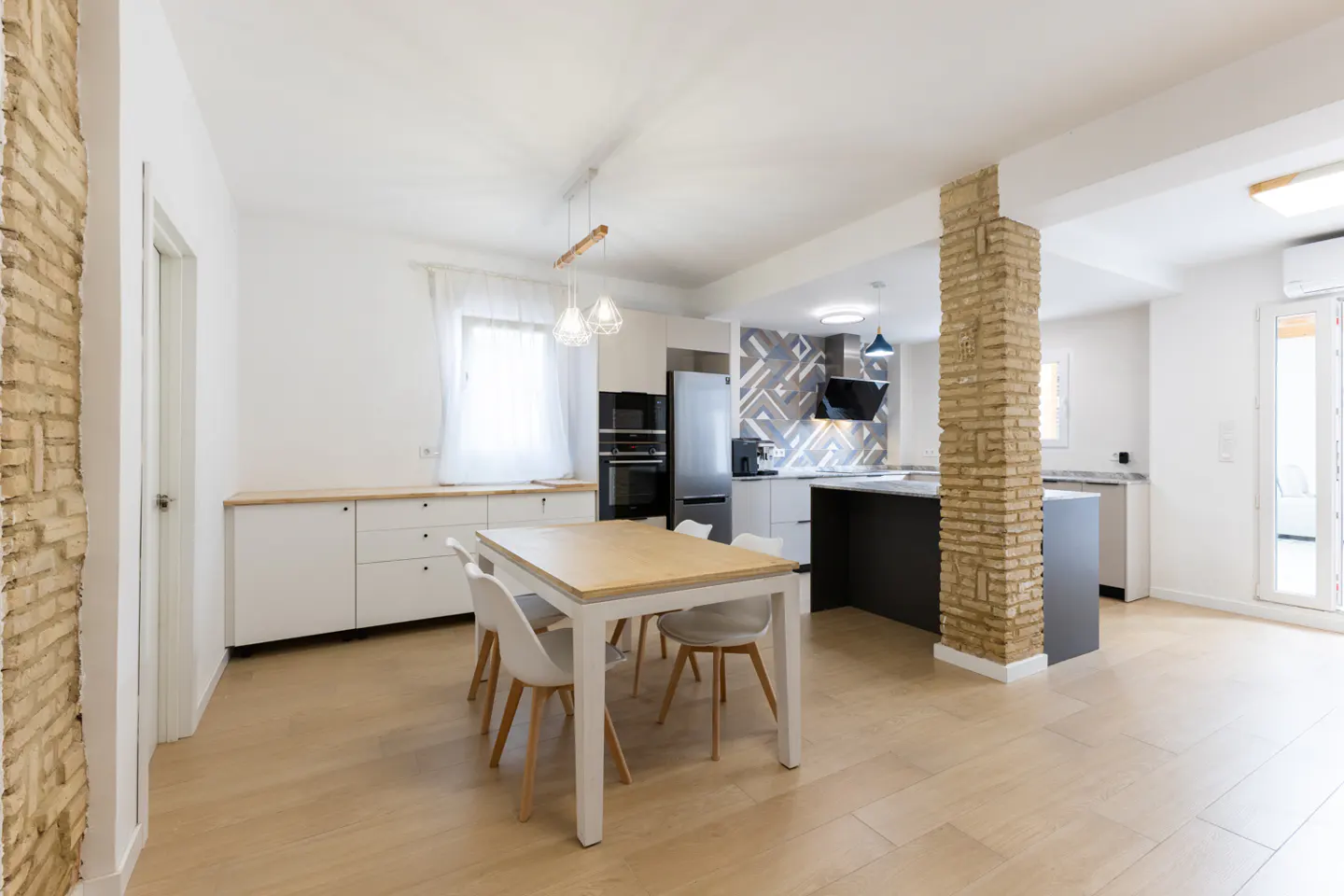 Open-concept kitchen and dining area with light wood floors, white walls, and exposed brick columns. A table with white chairs sits in the foreground.