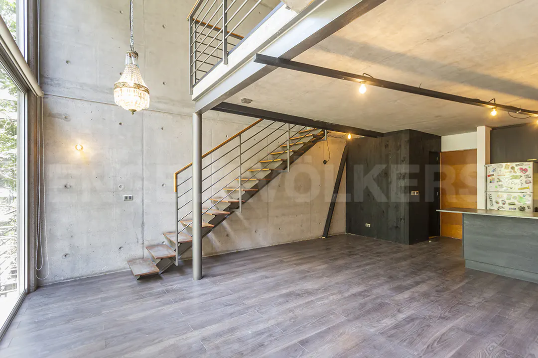 Interior view of a modern loft with concrete walls, wood floors, and a metal staircase leading to a mezzanine. A chandelier hangs from the ceiling.